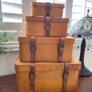 May include: Four wooden boxes with leather straps and buckles stacked on top of each other. The boxes are a light brown color and have a distressed look. The boxes are sitting on a wooden surface.