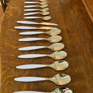 May include: A collection of gold-colored spoons and forks with white handles arranged on a wooden surface. The spoons have a rounded bowl shape, and the forks have four tines. The handles are a bright white color.