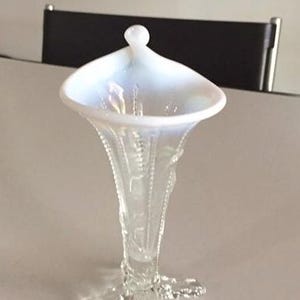 May include: A white iridescent glass vase with a flared top and a three-legged base. The vase has a delicate floral design.