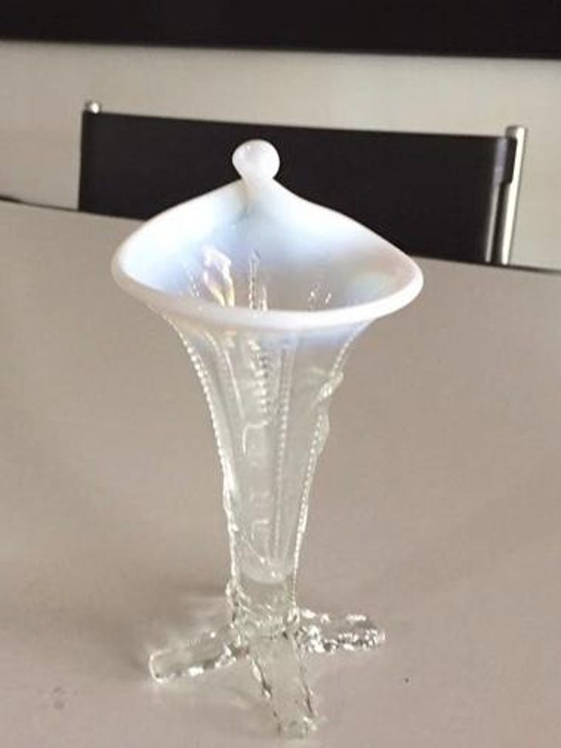 May include: A white iridescent glass vase with a flared top and a three-legged base. The vase has a delicate floral design.