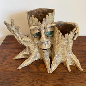 May include: A whimsical, tan-colored ceramic tree stump planter with a face and green eyes. The planter has two openings for plants and outstretched arms. The tree stump has a textured bark design and is sitting on a wooden surface.