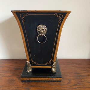 K&ouml;nnte beinhalten: Eine schwarz-goldene dekorative Box mit einem L&ouml;wenkopf-Ringgriff. Die Box hat eine trapezf&ouml;rmige Form mit goldenen Verzierungen und dekorativen Akzenten. Der Sockel hat verzierte F&uuml;&szlig;e und einen dekorativen Rand.