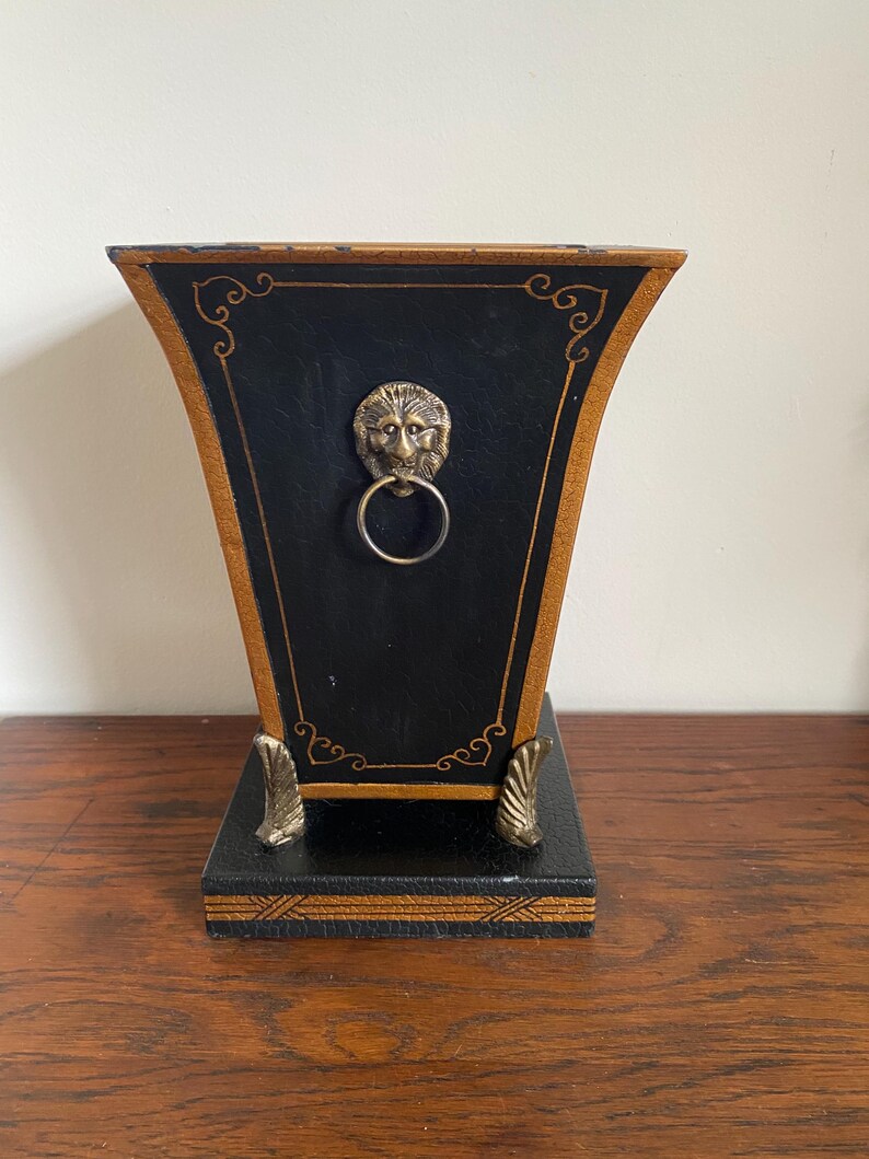 K&ouml;nnte beinhalten: Eine schwarz-goldene dekorative Box mit einem L&ouml;wenkopf-Ringgriff. Die Box hat eine trapezf&ouml;rmige Form mit goldenen Verzierungen und dekorativen Akzenten. Der Sockel hat verzierte F&uuml;&szlig;e und einen dekorativen Rand.