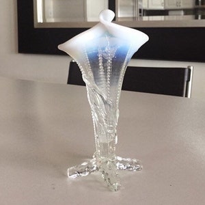 May include: A white and blue glass vase with a delicate floral design. The vase has a wide, flared opening and a slender, curved neck. The base of the vase is a four-legged stand.