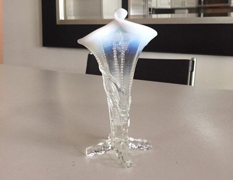 May include: A white and blue glass vase with a delicate floral design. The vase has a wide, flared opening and a slender, curved neck. The base of the vase is a four-legged stand.