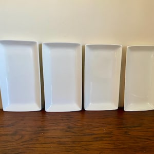May include: Four rectangular white ceramic serving platters of varying sizes are arranged on a dark wood surface. The platters have rounded corners and a smooth, glossy finish. They are displayed against a neutral wall.