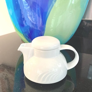 May include: White ceramic creamer or gravy boat with a lid and a wavy design. The creamer is on a black surface.