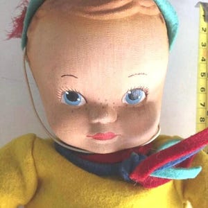 May include: A vintage cloth doll with blue eyes, a red and blue scarf, and a turquoise hat. The doll is wearing a yellow sweater.