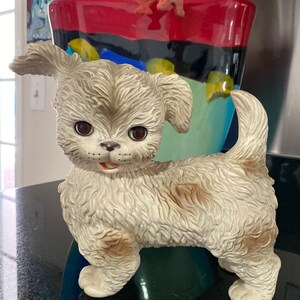 May include: A white ceramic figurine of a dog with brown markings. The dog has a fluffy texture and is standing with its head tilted to the side. The dog has large brown eyes and a playful expression.