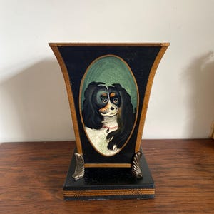 May include: A decorative wooden vase with a black exterior and a gold-colored trim. The vase features an oval portrait of a dog with black fur and a white face. The vase has decorative metal feet and sits on a black base.