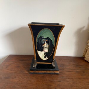 K&ouml;nnte beinhalten: Eine schwarz-goldene dekorative Vase mit einem gemalten Hundeportr&auml;t in einem ovalen Rahmen. Die Vase hat einen quadratischen Sockel mit dekorativen Metallf&uuml;&szlig;en und einen ausgestellten oberen Rand.