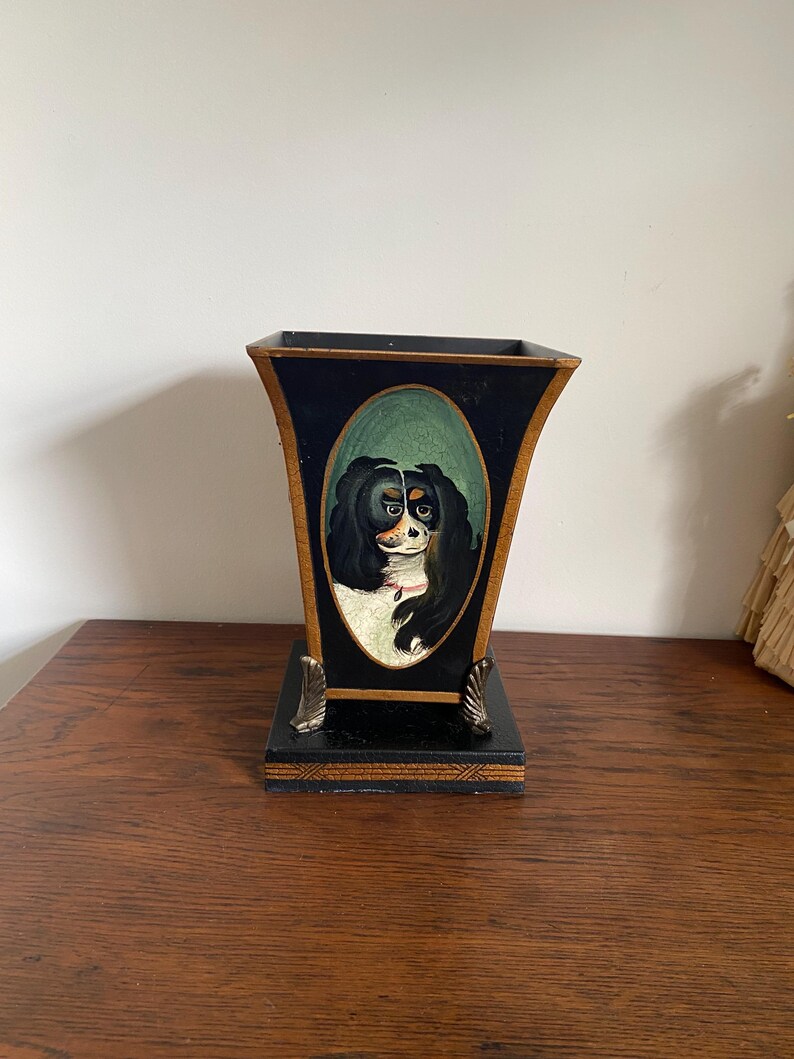 K&ouml;nnte beinhalten: Eine schwarz-goldene dekorative Vase mit einem gemalten Hundeportr&auml;t in einem ovalen Rahmen. Die Vase hat einen quadratischen Sockel mit dekorativen Metallf&uuml;&szlig;en und einen ausgestellten oberen Rand.