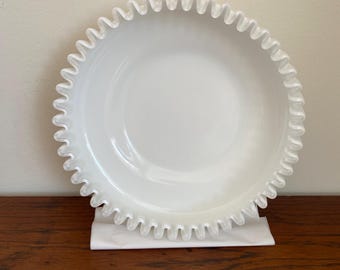 May include: A white, round glass bowl with a ruffled edge. The bowl is sitting on a white surface and is displayed on a wooden table. The bowl has a smooth, glossy finish.