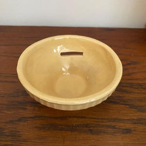 May include: A small, round, light yellow ceramic bowl with a slot on top. The bowl has a ribbed design around the edge and a smooth interior. This vintage-style bowl is likely a coin bank or a decorative piece.