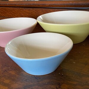 May include: Three ceramic bowls in pastel colors: pink, yellow, and blue. Each bowl has a white interior and a colored exterior. The bowls are arranged on a wooden surface, showcasing their angled design.