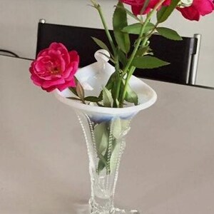 May include: A white milk glass vase with a scalloped rim and a delicate, ornate design. The vase has a tall, slender stem and a wide, flared base with three legs. The vase is filled with pink roses.