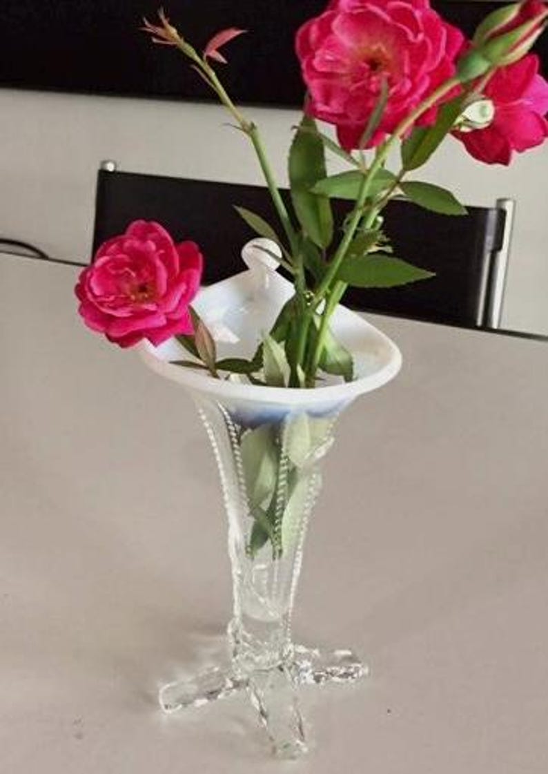 May include: A white milk glass vase with a scalloped rim and a delicate, ornate design. The vase has a tall, slender stem and a wide, flared base with three legs. The vase is filled with pink roses.