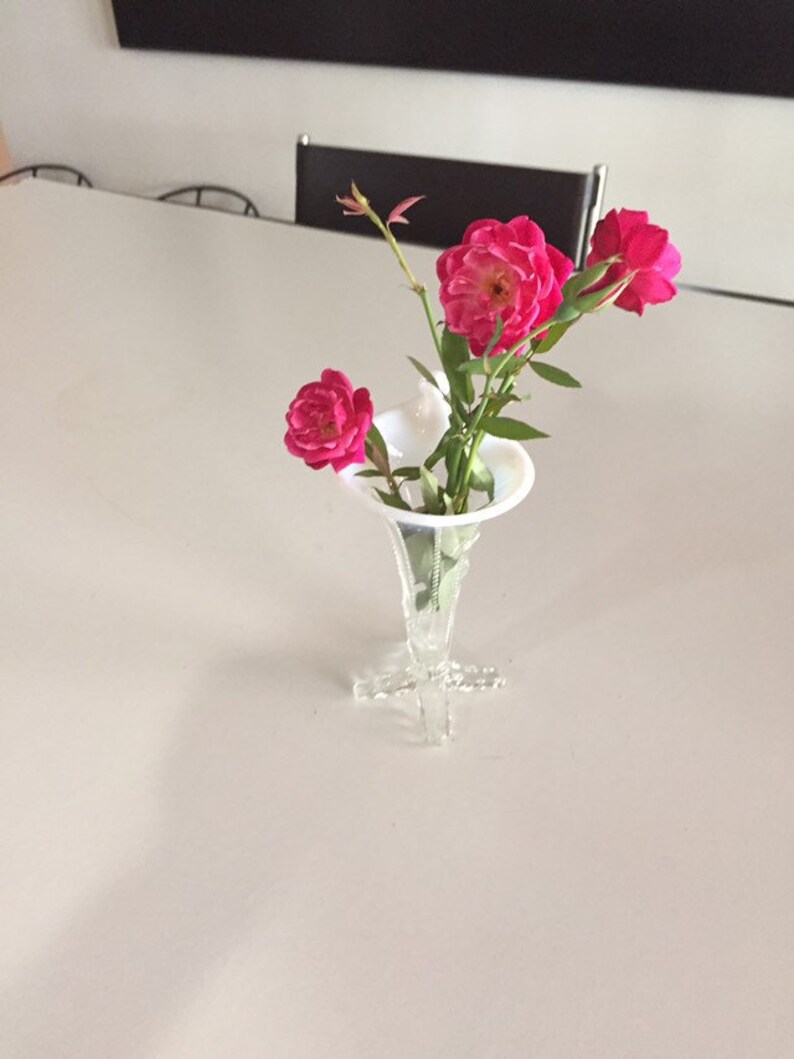 May include: A white glass vase with a flared opening and a delicate, intricate base holds a bouquet of pink roses.