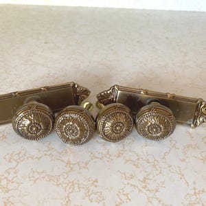 May include: Set of four antique brass door knobs with ornate details. Each knob features a raised, circular design with a central flower motif. The knobs are attached to two rectangular plates with decorative edges.