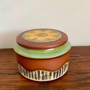 May include: A round ceramic lidded jar featuring a matte terracotta brown body with a green glazed rim. The lid displays a central yellow flower motif on a brown background. The base is decorated with a series of vertical black and white stripes.