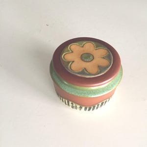 May include: A small ceramic jar with a lid featuring a stylized flower design. The jar is decorated with brown, green, and white stripes.
