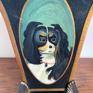K&ouml;nnte beinhalten: Dekorative Vase in Schwarz und Gold mit einem handgemalten Portr&auml;t eines Cavalier King Charles Spaniel-Hundes. Das Hundeportr&auml;t befindet sich in einem ovalen Rahmen mit rissigem, gealtertem Aussehen. Die Vase hat einen quadratischen Sockel und verzierte Metallf&uuml;&szlig;e.