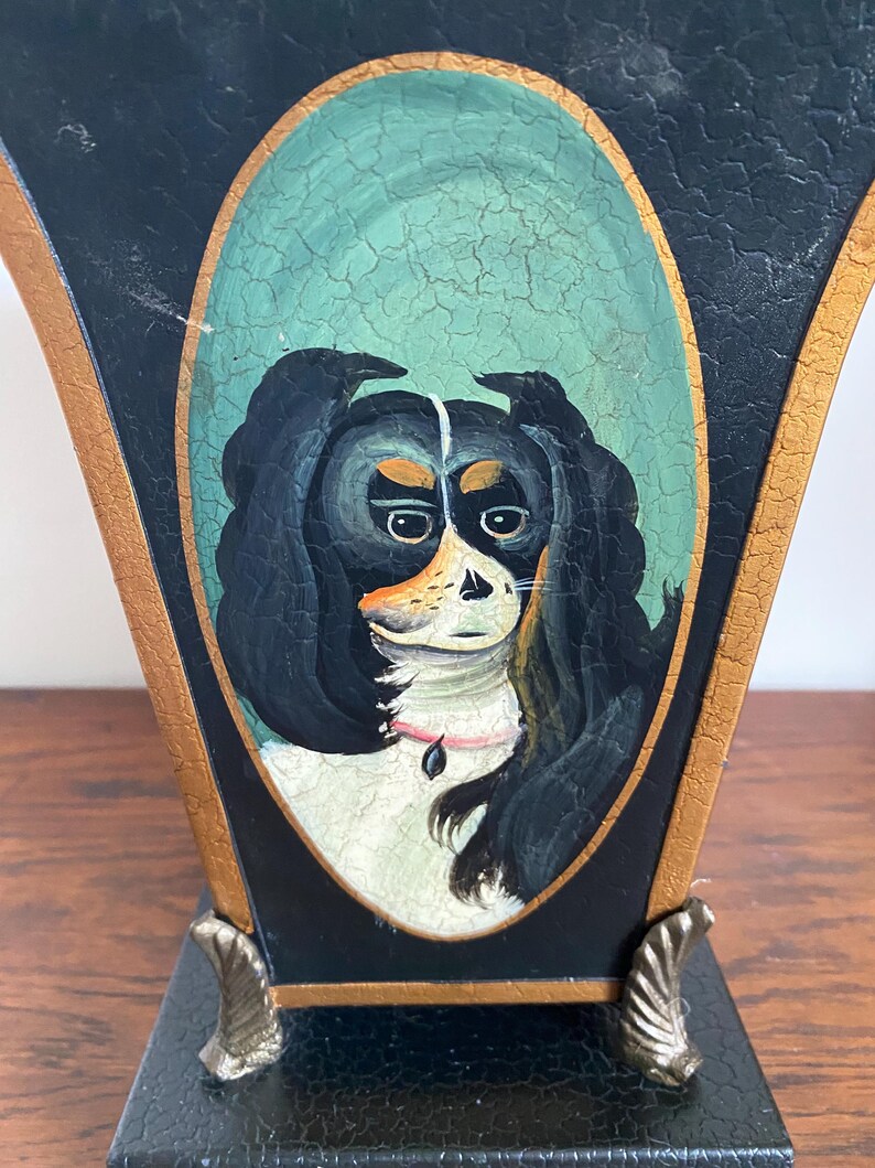 K&ouml;nnte beinhalten: Dekorative Vase in Schwarz und Gold mit einem handgemalten Portr&auml;t eines Cavalier King Charles Spaniel-Hundes. Das Hundeportr&auml;t befindet sich in einem ovalen Rahmen mit rissigem, gealtertem Aussehen. Die Vase hat einen quadratischen Sockel und verzierte Metallf&uuml;&szlig;e.