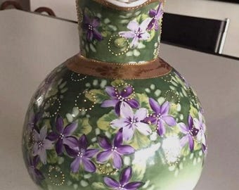 May include: A round ceramic pitcher with a green gradient base and a flared rim. The surface is decorated with hand-painted purple and light violet flowers, green leaves, and gold-colored dotted accents. It features a gold band around the neck.