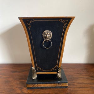 K&ouml;nnte beinhalten: Eine schwarz-goldene dekorative Box mit einem L&ouml;wenkopf-Ringgriff. Die Box hat eine trapezf&ouml;rmige Form mit goldenen Verzierungen und dekorativen Akzenten. Der Sockel ist quadratisch und ebenfalls schwarz und gold.