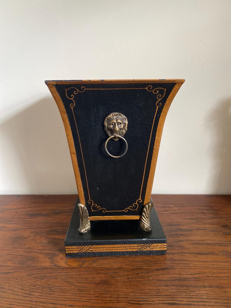 K&ouml;nnte beinhalten: Eine schwarz-goldene dekorative Box mit einem L&ouml;wenkopf-Ringgriff. Die Box hat eine trapezf&ouml;rmige Form mit goldenen Verzierungen und dekorativen Akzenten. Der Sockel ist quadratisch und ebenfalls schwarz und gold.