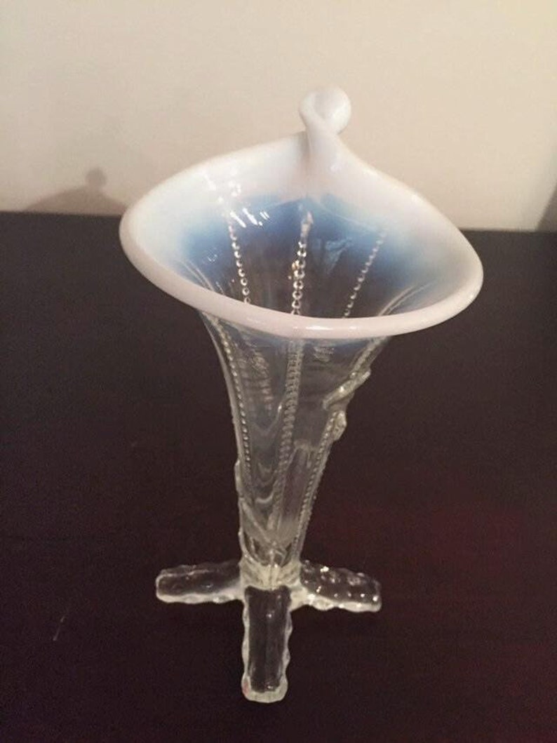May include: A clear glass vase with a white and blue opalescent top. The vase has a delicate, floral design and a flared opening. The base of the vase is a cross shape.