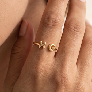Puede incluir: Un anillo abierto de tono dorado que presenta dos pequeñas letras tridimensionales redondeadas, H y G, situadas en cada extremo de la banda. El anillo tiene un acabado metálico liso y pulido, diseñado para llevarse en un dedo con espacio.