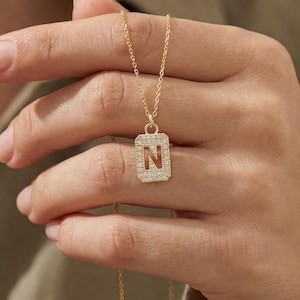 Op de afbeelding: Een goudkleurige ketting met een rechthoekige hanger met de letter "N" ingelegd met kleine, heldere kristallen. De ketting wordt in een hand gehouden, waarbij de fijne ketting en hanger worden benadrukt. De achtergrond is wazig.