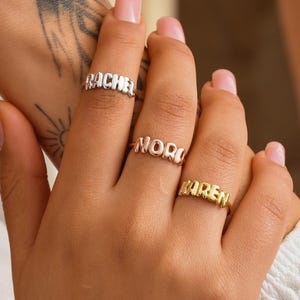 Könnte beinhalten: Drei gold- und silberfarbene Namensringe mit den Namen "RACHEL", "NORA" und "KAREN". Die Ringe werden an Fingern getragen, mit einem Arm und einer Hand im Vordergrund. Die Ringe sind im Fokus, mit unscharfem Hintergrund.