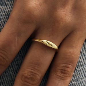 Könnte beinhalten: Ein goldfarbener Ring mit dem eingravierten Namen "Tony". Der Ring wird an einem Finger vor einem Jeans-Hintergrund getragen.