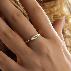 Könnte beinhalten: Ein goldfarbener Ring mit einem schlanken, länglichen, ovalen Mittelstück. Die Oberfläche des Ovals ist mit dem Namen Tony in einer klaren, dunklen, serifenlosen Schrift graviert. Der Ring ist dünn und glatt und hat ein gleichmäßiges Finish.
