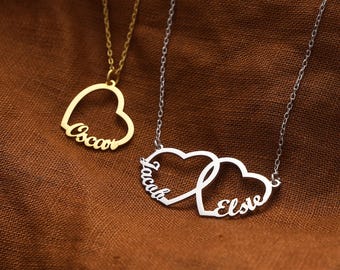 Op de afbeelding: Twee kettingen met hartvormige hangers. Eén is goudkleurig met de naam "Oscar" gegraveerd. De andere is zilverkleurig, met twee in elkaar grijpende harten met de namen "Jacob" en "Elsie". Beide kettingen liggen op een bruine achtergrond.