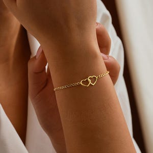 Op de afbeelding: Een goudkleurige armband met twee hartvormige bedels. De armband heeft een delicaat kettingontwerp. De afbeelding toont de armband om een pols. De achtergrond is een zachte, neutrale kleur.