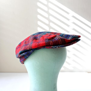 May include: A red, blue, and green plaid flat cap. The cap is displayed on a light blue mannequin head. The cap has a classic, rounded shape with a small brim. The plaid pattern is a traditional design.