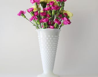 Op de afbeelding: Een witte hobnail vaas bevat een boeket van roze en gele anjers. De vaas heeft een uitlopende bovenkant en een brede basis. De bloemen staan in volle bloei, met groene stelen en bladeren.