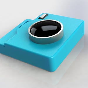 May include: A blue plastic camera icon with a silver lens. The camera is a 3D design and is sitting on a white surface.