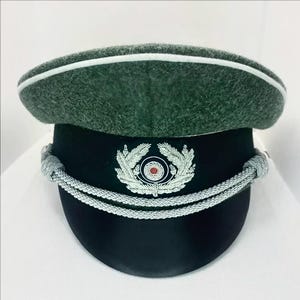 May include: A dark green military hat with a black visor and a silver emblem. The hat features a white band around the top and silver cords. The emblem is a detailed design with a red centre.