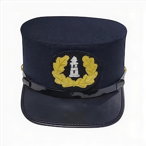 Könnte beinhalten: Eine marineblaue Mütze mit schwarzem Schirm und einem kreisförmigen Emblem mit einem Leuchtturm und goldenen Lorbeerblättern. Die Mütze hat eine zylindrische Krone und ein glänzendes schwarzes Band. Goldene Akzente sind an den Seiten sichtbar.
