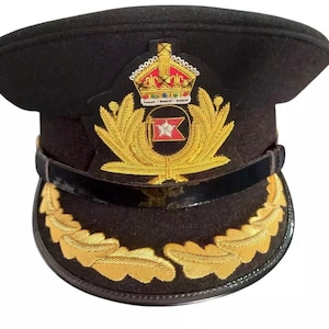 Könnte beinhalten: Schwarze Militärmütze mit goldenem Emblem mit Krone, Lorbeerblättern und rotem und weißem Wappen. Die Mütze hat eine schwarze Krempe und ein schwarzes Band am unteren Rand.