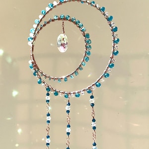 May include: A hanging ornament featuring two concentric copper wire circles adorned with small turquoise and clear beads. A single iridescent crystal hangs in the center, while three beaded strands with teardrop-shaped crystal pendants hang below.