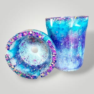 May include: A resin ashtray and a matching cup featuring a swirling blend of blue, purple, and white colors. Both items are embedded with multi-colored glitter particles throughout the surface, creating a vibrant, reflective, and textured appearance.