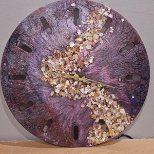 May include: A circular wall clock featuring a swirling purple and black resin finish with a central diagonal band of crushed iridescent seashells. The clock face has twelve elongated rectangular cutouts for hour markers and gold-colored hands.