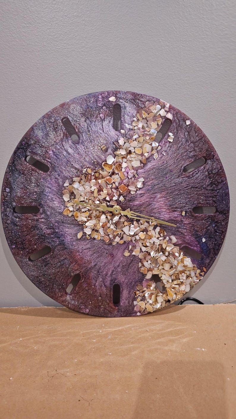 May include: A circular wall clock featuring a swirling purple and black resin finish with a central diagonal band of crushed iridescent seashells. The clock face has twelve elongated rectangular cutouts for hour markers and gold-colored hands.