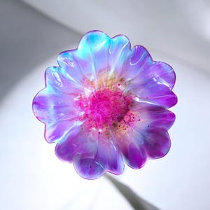 May include: A flower-shaped decorative object featuring translucent petals in shades of iridescent blue, purple, and white. The center of the flower is a vibrant, saturated magenta with textured, darker speckles throughout the core.
