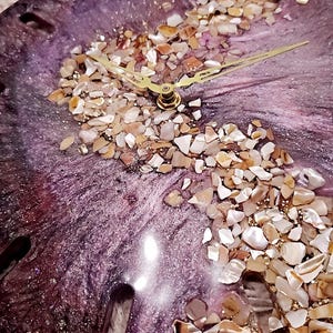 May include: A circular wall clock featuring a deep purple and metallic swirl resin base. An S-shaped band of crushed iridescent seashells and gold glitter runs across the center. The clock has thin gold-toned metal hour and minute hands.
